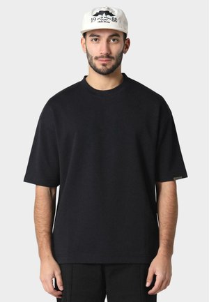 Svart oversized t-skjorte med korte ermer, rund halslinning og subtil detalj med sideetikett, laget av teksturert bomullsstoff.