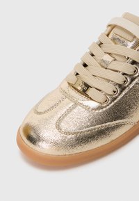 Sneaker metallico dorato con una finitura liscia e testurizzata, lacci beige e un sottile accento di logo sulla linguetta. Suola in gomma.