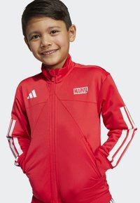 Rode ritsjack met een Marvel-logo, gemaakt van gladde stof met witte accenten en drie strepen op elke mouw. Inclusief zakken.