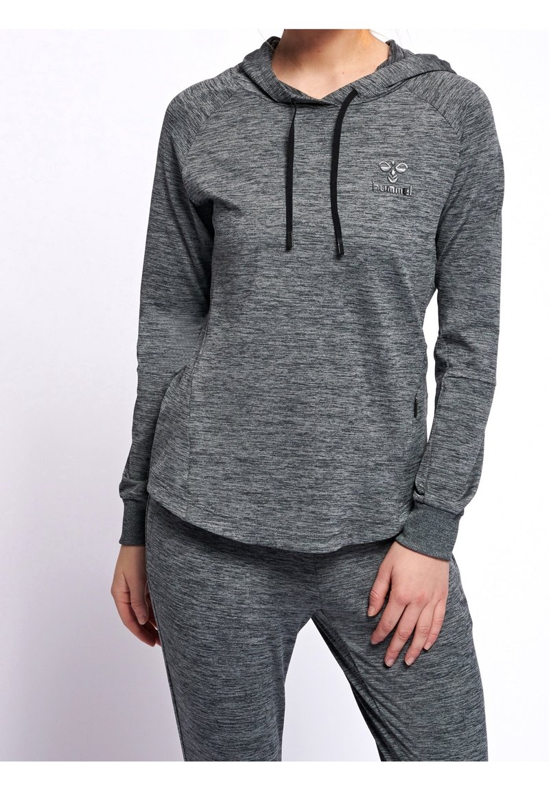 Femme portant un sweat à capuche gris assorti à manches longues et un pantalon avec des cordons noirs, debout avec une main reposant sur sa cuisse.