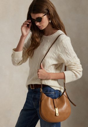 Moteris dėvi kreminį megztinį su pynėmis ir mėlynas džinsus, taiso vėžlio rėmelio saulės akinius, laiko rudą odinę peties rankinę su aukso spalvos sagtimi.