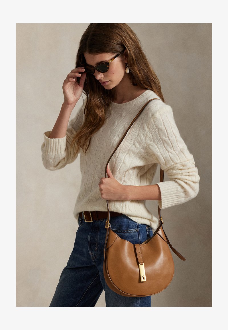 Naine kandes kreemikat palmikutega kampsunit ja siniseid teksaseid, kohandades kilpkonnamustriga päikeseprille, hoides pruuni nahast õlakotti kullast kinnitusdetailiga.