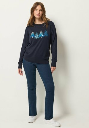 Femme portant un pull bleu marine avec des motifs d’arbres bleus et argentés, un jean bleu foncé et des baskets blanches, debout devant un fond clair uni.