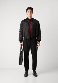 Zwarte bomberjack met drukkers, bordeauxrood t-shirt, zwarte joggingbroek met logo, zwarte schoenen en een grijze totebag. Zachte texturen overal.