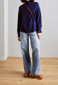 Mörkblå turtleneck-tröja i kombination med ljusblå vida jeans och tanfärgade träklackar med silvernitar; står på ett ljust trägolv.