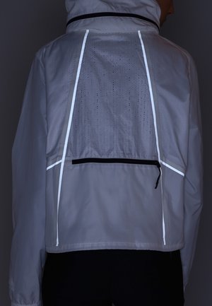 Osoba ubrana w szarą kurtkę z odblaskowymi paskami i perforowanym panelem wentylacyjnym na górnej części pleców, stojąca na tle ciemnego tła.