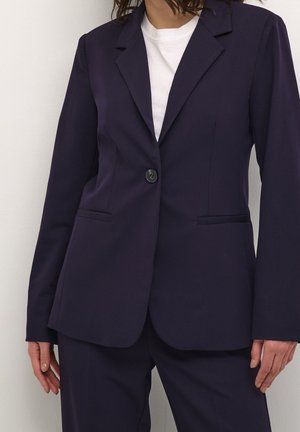 Femme portant un blazer bleu marine foncé à un bouton et un pantalon assorti sur un t-shirt blanc à col rond, debout devant un fond blanc uni.