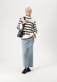 Melnbaltā svītrainā adīta turtlenecka džemperis, gaiši zilas denima maxi svārki, melnas kurpes, rokā tur mazu melnu somiņu.