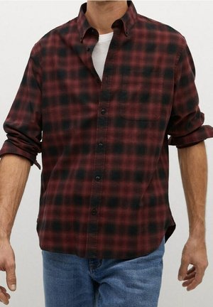 Mann trägt ein rot-schwarz kariertes Langarmhemd über einem weißen T-Shirt mit hochgekrempelten Ärmeln und blauer Jeans.