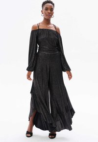 Zwarte, off-the-shoulder jumpsuit met lange mouwen, glinsterende stof, wijduitlopende pijpen en gerokte accenten langs de zoom.