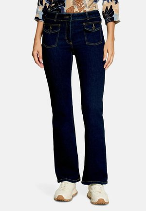 Femme portant un jean denim bleu foncé avec des poches à rabat à l’avant, un haut à motif floral et des baskets blanches, debout devant un fond uni.