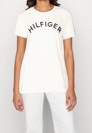 Kobieta nosząca białą, krótką koszulkę z nadrukiem "HILFIGER" na czarno na piersi oraz białe spodnie.