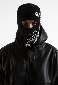Balaclava nera lavorata a maglia con design di scorpione bianco. Accompagnata da una giacca nera in pelle con zip, dalla texture liscia e con coulisse regolabili.