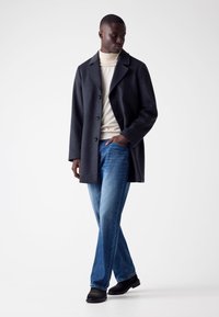 Cappotto in lana grigio scuro con ampi rever, chiusura con bottoni; dolcevita crema, jeans in denim blu, scarpe casual nere; silhouette slim fit.