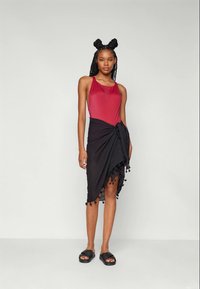 Κόκκινο μαγιό χωρίς μανίκια, μαύρη φούστα τύπου wrap με φούντες, μαύρα επίπεδα σανδάλια. Ομαλή υφή, ρέον ύφασμα και γωνιώδης πτυχώσεις.