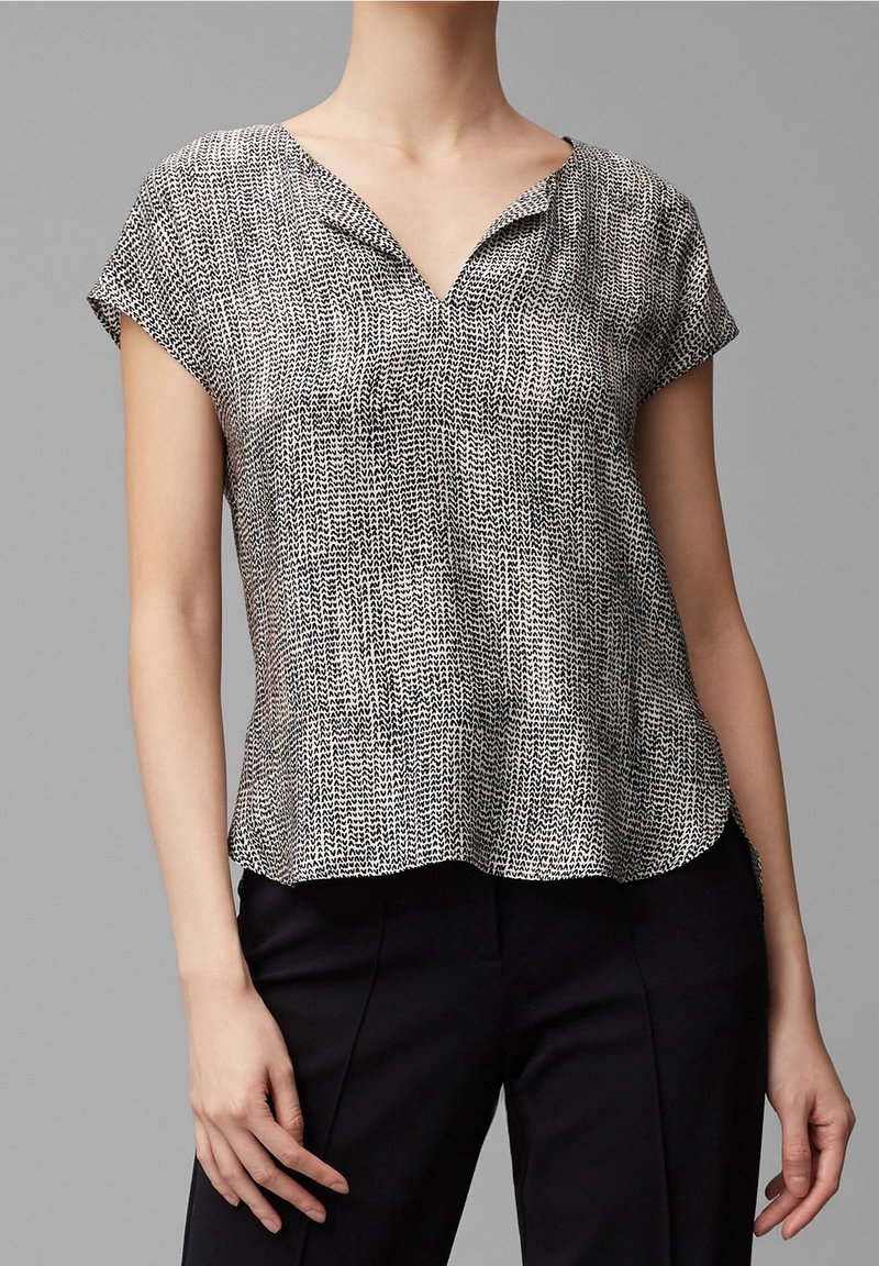 Blouse à manches courtes avec un motif noir et blanc, présentant un décolleté en V, une texture douce et un design aérien, portée avec un pantalon noir.
