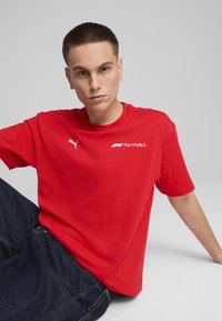 Rød bomulds T-shirt med rund hals, korte ærmer og hvide Puma- og Formel 1-logoer på forsiden. Glat tekstur, afslappet pasform.