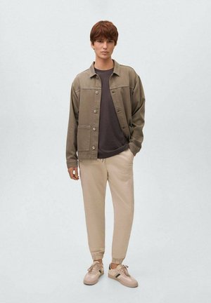 Personne debout portant une veste olive, une chemise gris foncé, un pantalon de jogging beige et des baskets beiges avec des rayures noires sur un fond clair.