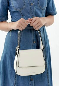 Bolso blanco con una textura suave, acentos negros, forma rectangular y un asa corta y brillante, sostenido por una persona vestida con un vestido de mezclilla.