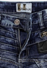 Jeans in denim scuro con zip, chiusura a bottone metallico e cuciture gialle a contrasto. Include una toppa in pelle nera sul retro.