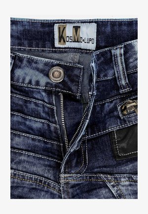 Donkerdenim jeans met een ritssluiting, een metalen knoopsluiting en contrasterende gele stiksels. Bevat een zwart leren patch op de achterkant.