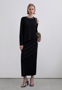 Mujer con un vestido negro asimétrico con un recorte en un hombro, sosteniendo un clutch dorado y calzando sandalias de tacón alto plateadas.