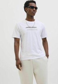Hvid bomulds t-shirt med "JACK & JONES" trykt i grå. Iført lyse beige bukser viser modellen en afslappet pasform og korte ærmer.