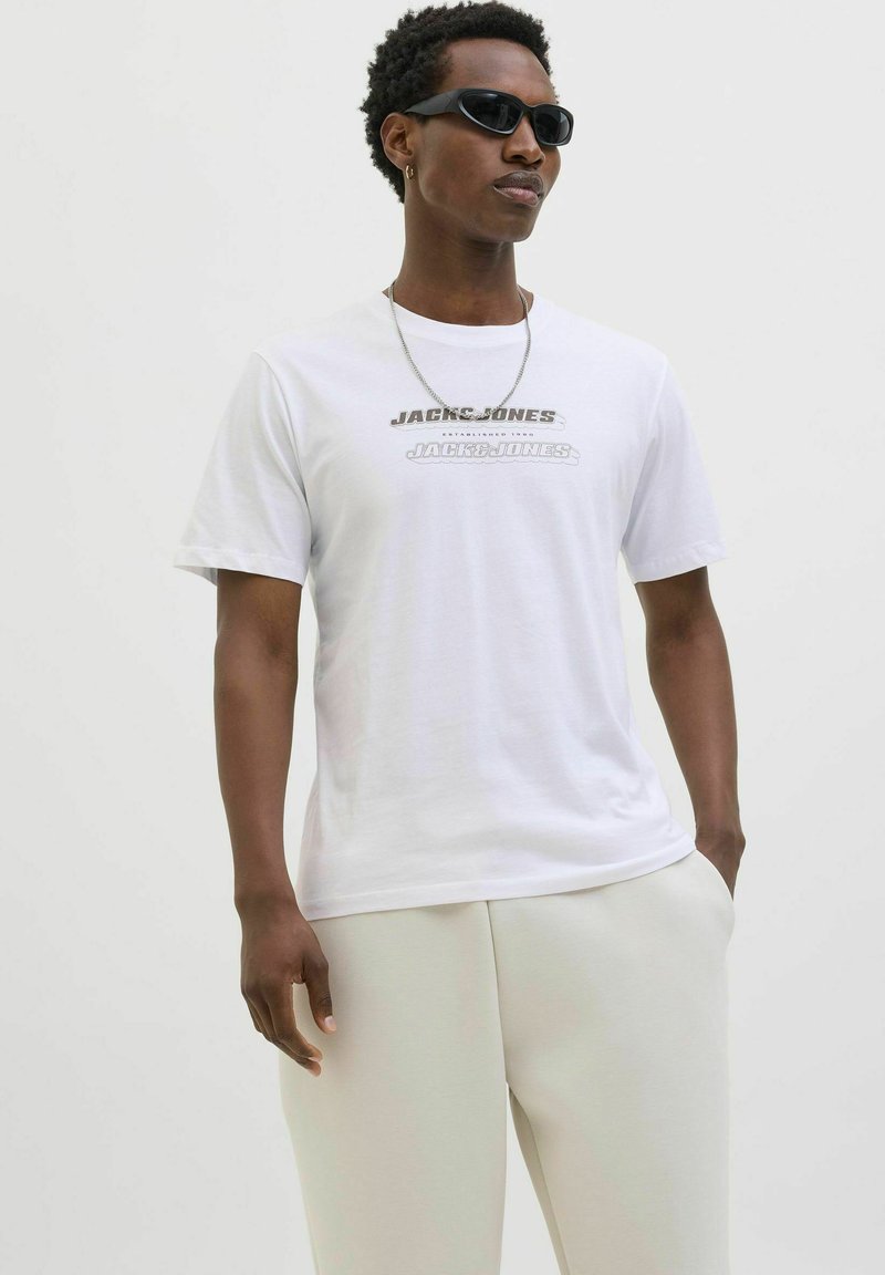 Hvid bomulds t-shirt med "JACK & JONES" trykt i grå. Iført lyse beige bukser viser modellen en afslappet pasform og korte ærmer.