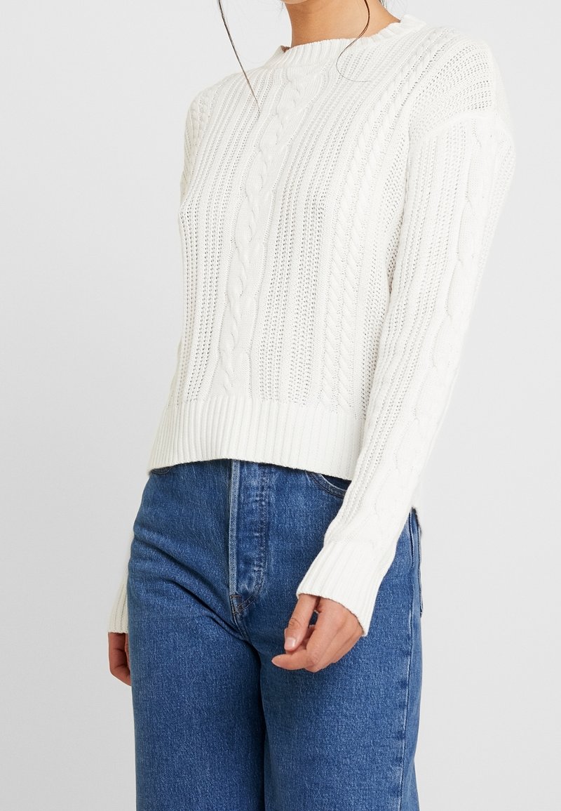 Woman wearing a white cable-knit sweater with textured patterns and blue high-waisted denim jeans against a plain background.