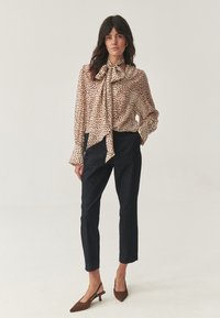 Femme portant un chemisier beige à pois avec un grand nœud, un pantalon court sombre et des escarpins à talons kitten pointus marron, se tenant devant un fond uni.