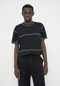 Μαύρο μπλουζάκι με αντίθετη γκρι ραφή, κοντά μανίκια και στρογγυλή λαιμόκοψη. Συνδυασμένο με μαύρο παντελόνι φόρμας με κορδόνι στη μέση. Απλός, μονόχρωμος σχεδιασμός.