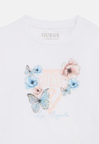Λευκό βαμβακερό T-shirt με γραφικά από λουλούδια και πεταλούδες σε μπλε και ροζ, με το κείμενο "GUESS?" και "Los Angeles" σε απαλό πορτοκαλί χρώμα.