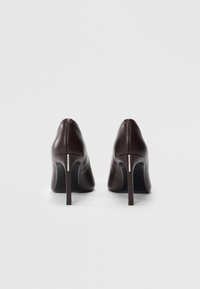 Un paio di scarpe con tacco alto in pelle marrone scuro, con tacchi a stiletto sottili, fotografate di spalle su uno sfondo bianco.