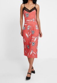 Femme portant une robe midi sans manches corail avec imprimé floral noir et encolure en dentelle, assortie à des escarpins noirs �à bout pointu avec des brides à la cheville.