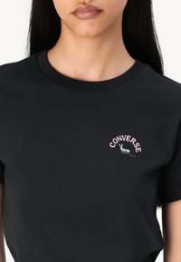 T-shirt preta de algodão com decote redondo e mangas curtas. O design da frente inclui o texto "CONVERSE" em rosa com um gráfico de formiga.