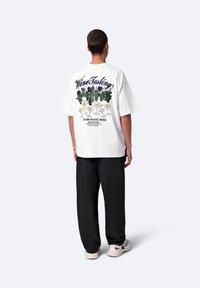 Osoba z krótkimi włosami ubrana w biały oversize'owy t-shirt z ilustracjami winorośli i kieliszka wina oraz czarne spodnie, stojąca tyłem.