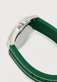 Hõbe kellakorpus, millel on tekstuuriga serv ja roheline kummist rihm, vaadatuna külgvaates, rõhutades kroon ning rihma detaile.