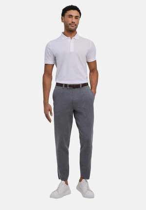 Mann steht mit den Händen in den Taschen, trägt ein weißes Poloshirt, graue Hose, braunen Gürtel und weiße Sneakers vor einfarbigem Hintergrund.