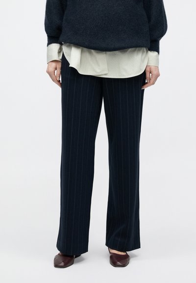 Marineblaue Nadelstreifenhose in gerader Schnittführung, gefertigt aus einem glatten Stoff, kombiniert mit einem dunklen, strukturierten Pullover und einem hellen Hemd darunter.