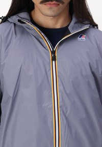 Veste imperméable grise avec une fermeture éclair présentant des rayures jaunes, blanches et noires. Comprend une capuche et le logo K-Way sur le haut gauche de la poitrine.