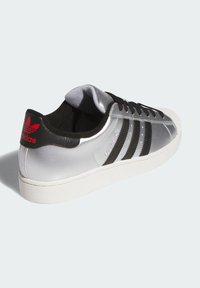 Baskets synthétiques argentées avec des accents noirs, trois bandes noires sur le côté et un logo rouge sur le talon. Elles possèdent une semelle en caoutchouc blanche.