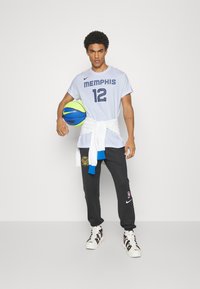 T-shirt gris clair avec "MEMPHIS 12" en navy, pantalon de survêtement noir avec logo, et baskets blanches. Tenant un ballon de basketball multicolore.