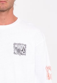 Biała, długiakowiec T-shirt z czarnym wzorem na klatce piersiowej, który ma skomplikowane desenie, oraz pomarańczowym akcentem graficznym na prawym rękawie.