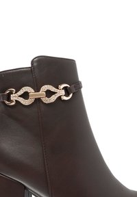 Stivale alla caviglia in pelle marrone con tacco spesso, accentuato da una catena decorativa con strass e una texture elegante e liscia.