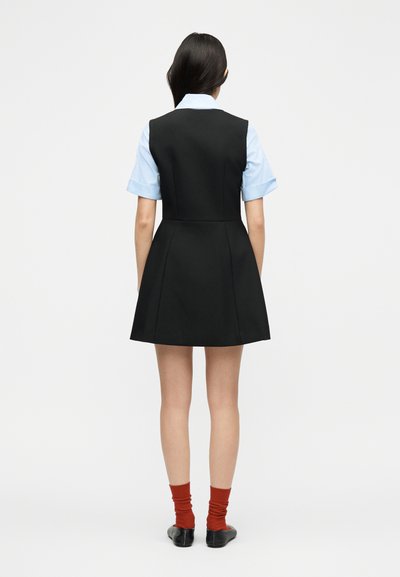 Γυναίκα που στέκεται με την πλάτη προς το φακό, φοράει μαύρο αμάνικο φόρεμα πάνω από γαλάζιο κοντομάνικο πουκάμισο, κόκκινες κάλτσες και μαύρα παπούτσια σε απλό φόντο.