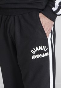 Sorte joggingbukser lavet af blødt stof, med hvide sidelister og et logo print "GIANNI KAVANAGH" på venstre lår. Designet til komfort.