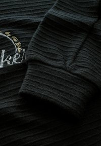 Schwarz geripptes Gewebe, das eine Nahaufnahme des Bündchens eines Sweatshirts zeigt, mit gesticktem Text in metallischem Gold und weißen Akzenten.