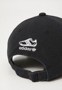 Juodas beisbolo kepurė su baltu siuvinėtu Adidas sportbačių ir logotipo dizainu, su išlenktu kraštu ir reguliuojama juostele gale.