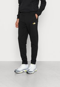 Černé tepláky se slim fit, vyrobené z měkké látky, s žlutým logem na levém stehně a bočními kapsami. Nosit s šedými a žlutými teniskami.
