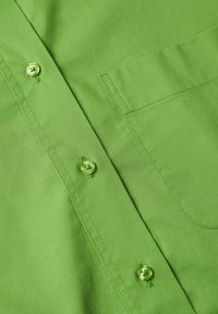 Gros plan sur le devant d'une chemise verte montrant trois boutons et une poche cousue sur un tissu lisse.